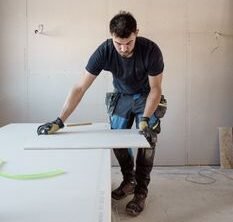 A blue collar worker measuring a plank indoors in a bright workshop.